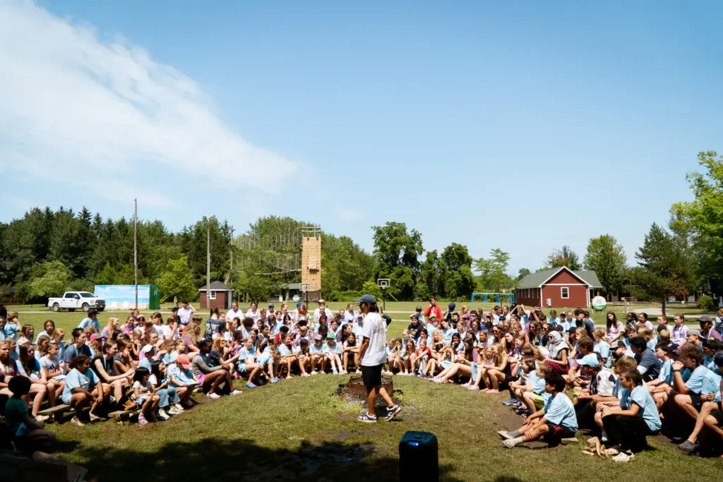 A summer camp field with kids gathered around a camp counsellor.